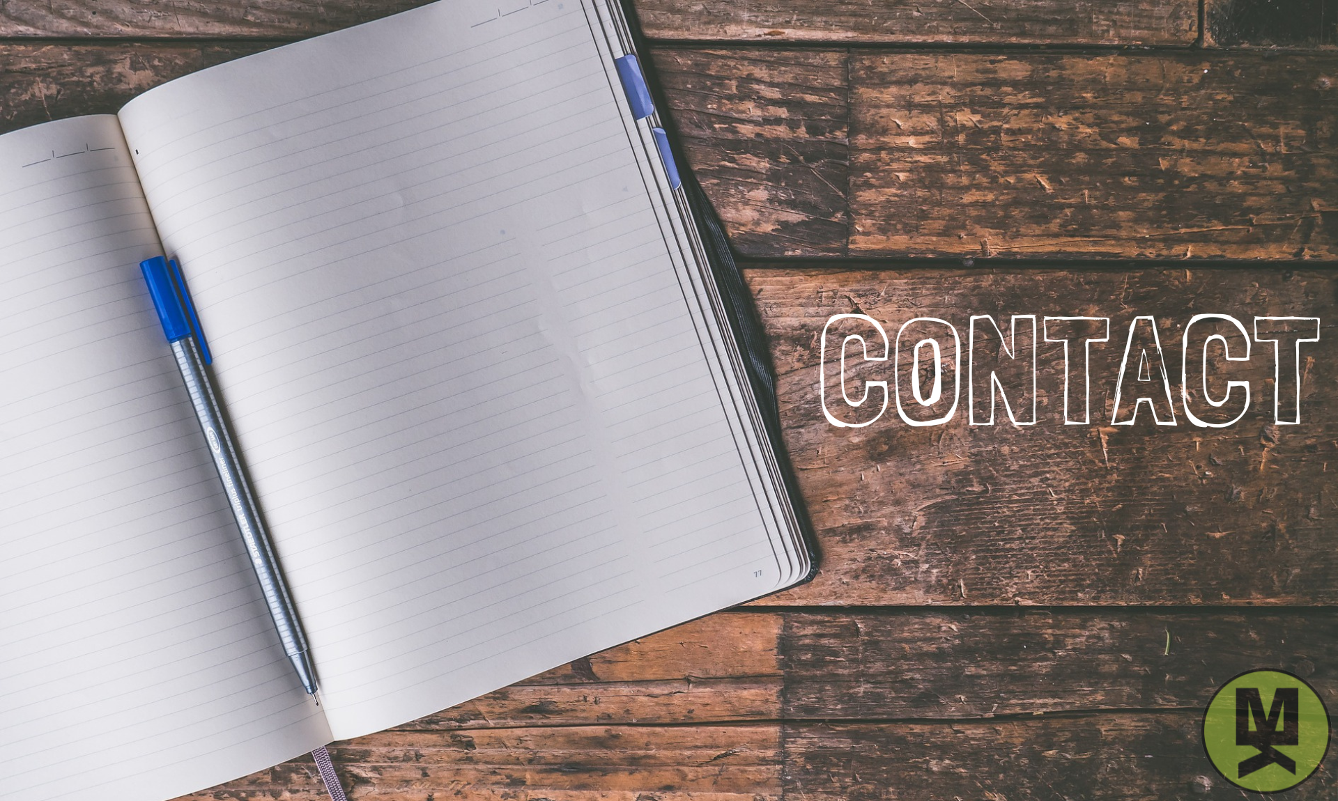 An open copy book with a pen in the middle of the book as it rests on a wooden table. The word "Contact" is beside the book.