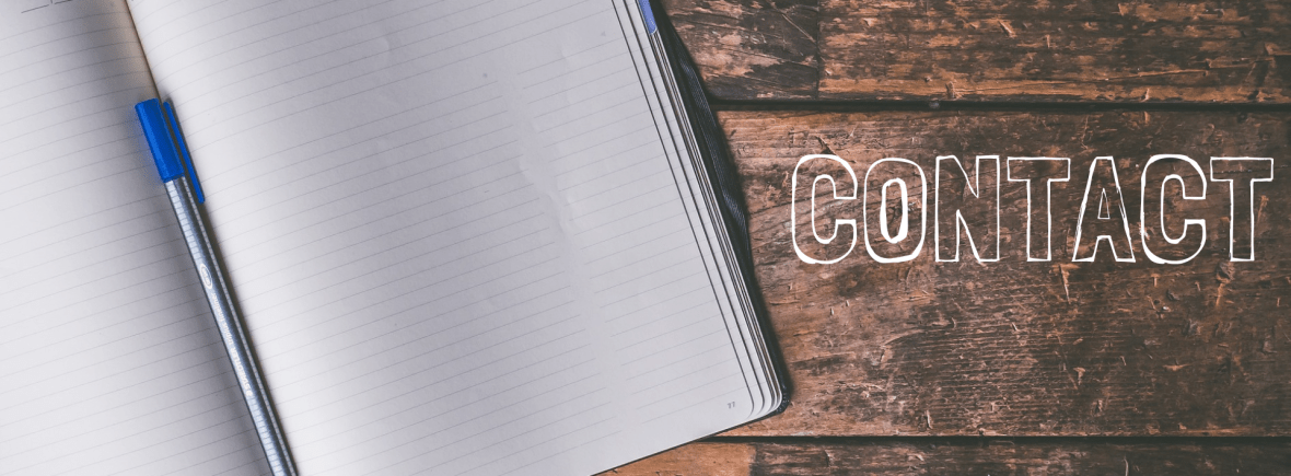An open copy book with a pen in the middle of the book as it rests on a wooden table. The word "Contact" is beside the book.
