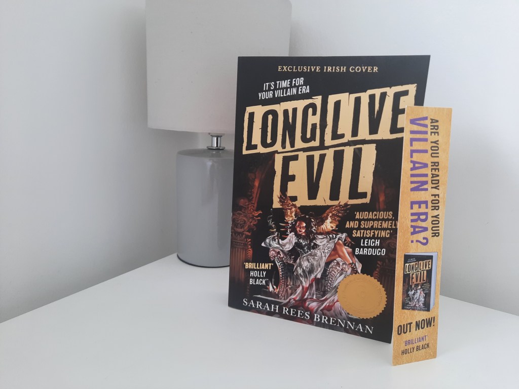 A copy of Long Live Evil by Sarah Rees Brennan sits on a bedside locker next to a grey lamp. Propped against the book is a promotional bookmark stating "Are you ready for your villain era?"