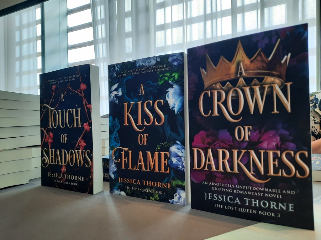 The Lost Queen book trilogy standing on a table at Octocon 2024 by Jessica Thorne. A Touch of Shadows, A Kiss of Flame, and A Crown of Darkness.